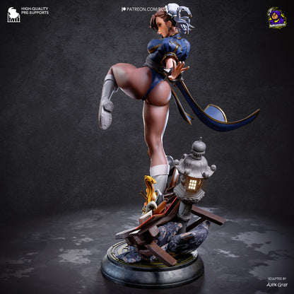 A detailed collectible statue of Chun-Li in a dynamic kicking pose, mounted on a decorative base with a lantern and rocks. The figure wears a blue and white outfit with gray boots and headpiece.
