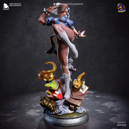 A highly detailed collectible statue of Chun-Li in her signature martial arts pose, standing on a circular base with miniature props including a green frog, a banana, and a skull. The statue is set against a dark, textured studio background.